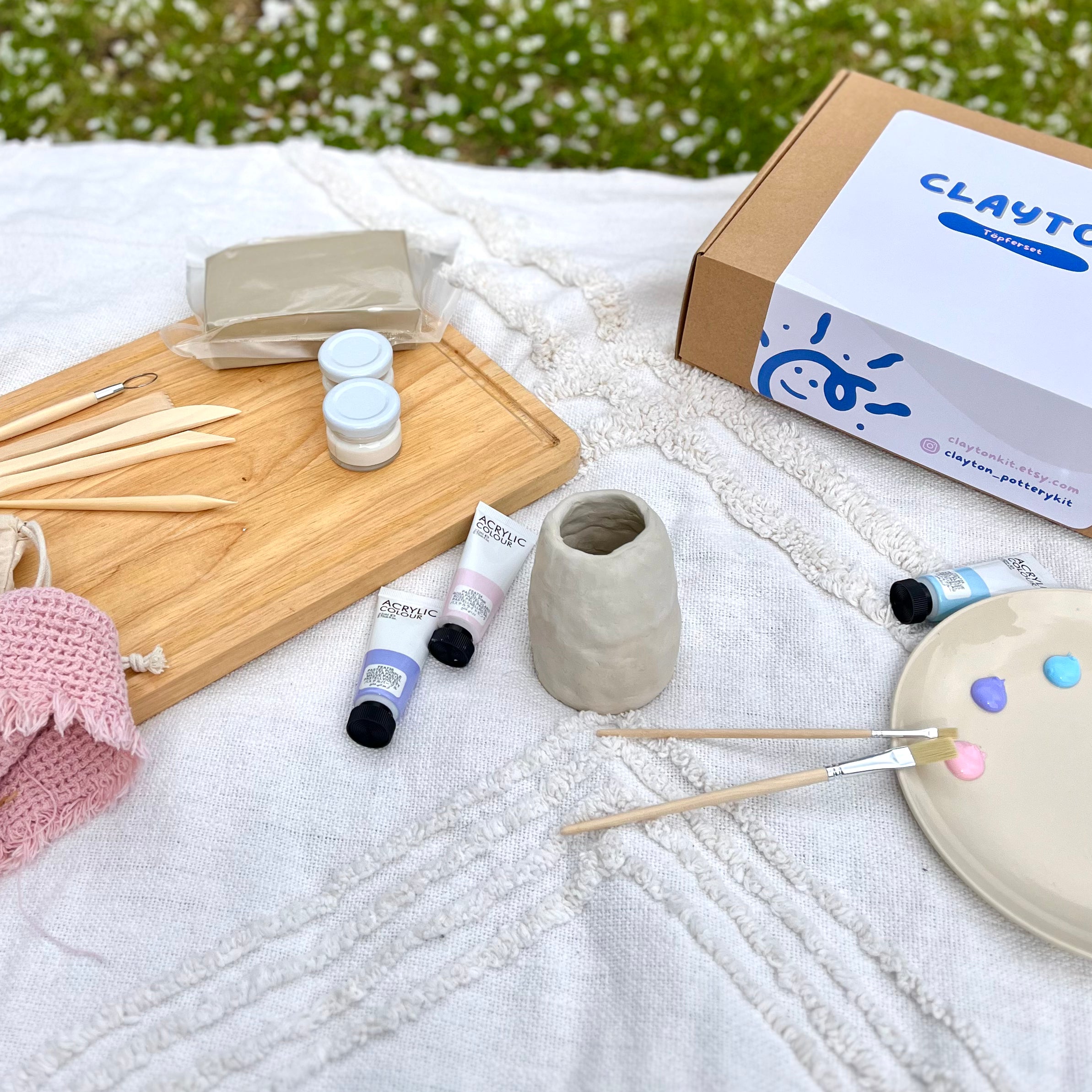 Ein Töpferset auf einer Picknickdecke mit Ton, Farben und Modellierwerkezuegen. Im Hintergrund ein Töpferset.